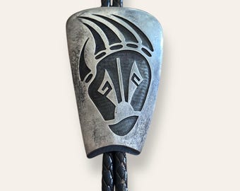 May include: A silver-toned bolo tie with a bear claw design. The pendant features a detailed bear claw and face engraving. The black braided leather cord is visible, creating a classic Western accessory.