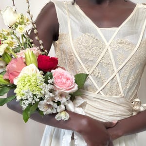 May include: A person wearing a white sleeveless dress with lace detailing and a white sash. They are holding a bouquet of flowers, including pink roses, white lilies, and green hydrangeas.