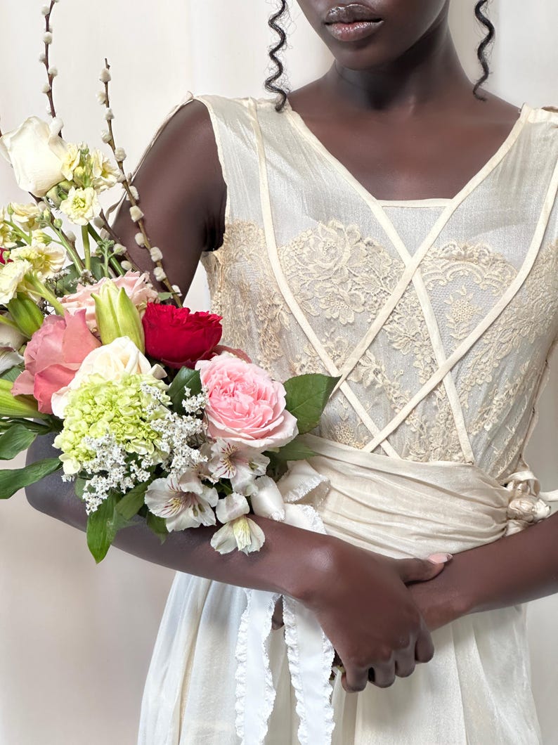 May include: A person wearing a white sleeveless dress with lace detailing and a white sash. They are holding a bouquet of flowers, including pink roses, white lilies, and green hydrangeas.