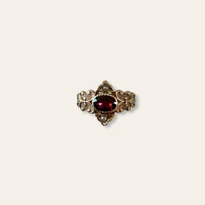 May include: A gold-toned ring featuring a large, oval, deep red gemstone at its center. The ring has ornate filigree detailing and small, round, white gemstones on either side of the central stone. The ring's design is symmetrical and elegant.
