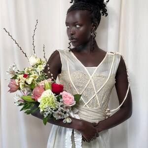 May include: A person wearing a white sleeveless dress with lace detailing and a white sash. They are holding a bouquet of flowers that includes pink, white, red, and green flowers.