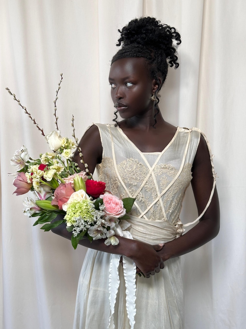 May include: A person wearing a white sleeveless dress with lace detailing and a white sash. They are holding a bouquet of flowers that includes pink, white, red, and green flowers.