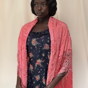 May include: A woman wearing a coral-colored shawl with intricate floral embroidery and fringe. The shawl is draped over a dark blue floral dress. The dress has a ruffled neckline and a pattern of colorful flowers.