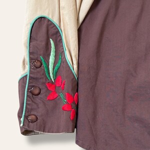 May include: Close-up of a western-style shirt sleeve. The sleeve is a combination of tan and brown fabric. The cuff features a brown embroidered design with red flowers, green leaves, and a teal border. Three brown buttons secure the cuff.