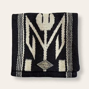 May include: Folded black and white woven textile with a geometric design. The pattern features a central stylized floral motif and vertical stripes. A small, silver-colored metal embellishment is visible on the front.