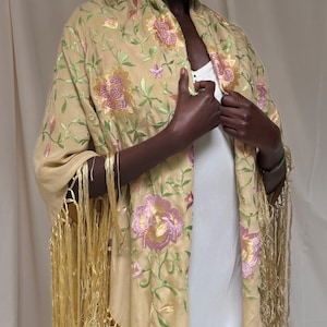 May include: A beige shawl with floral embroidery in pink, green, and gold. The shawl features long, gold fringe along the edges. The floral design includes large flowers and smaller blossoms with green leaves. The shawl is draped over the shoulders.