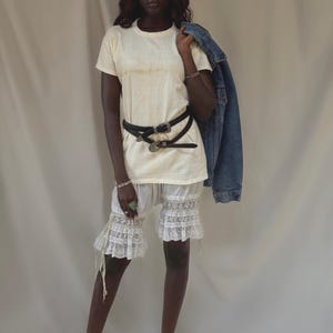 May include: A cream-colored short-sleeve t-shirt paired with white lace-trimmed bloomers, two black leather belts with silver buckles, and a blue denim jacket draped over one shoulder. The ensemble features a layered look with textured lace.