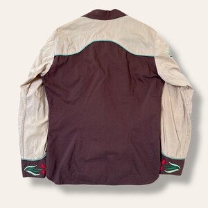 May include: Back view of a western-style shirt. The shirt features a brown body with tan sleeves and yoke. The design includes turquoise trim and red floral embroidery on the cuffs.