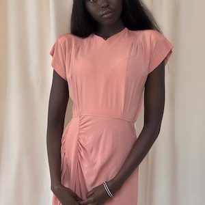 May include: A peach-colored dress with short sleeves and a gathered detail on the side. The dress has a fitted waist and a flowing skirt. A silver bracelet with red and white accents is visible on the wrist.