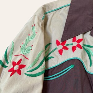 May include: Tan and brown western-style shirt with embroidered floral and cactus designs. The shirt features red flowers, green leaves, and cacti, with turquoise trim. The collar is brown, and the sleeves are tan.
