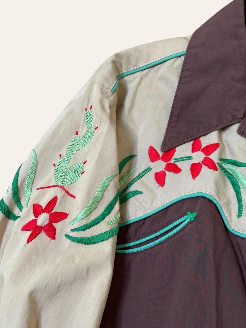 May include: Tan and brown western-style shirt with embroidered floral and cactus designs. The shirt features red flowers, green leaves, and cacti, with turquoise trim. The collar is brown, and the sleeves are tan.
