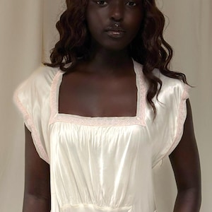 May include: Ivory-colored satin nightgown with a square neckline and cap sleeves. The neckline and sleeve edges are trimmed with a delicate pink Greek key pattern. The gown has a gathered waist and a flowing skirt.