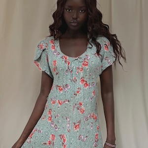 May include: A light green dress with a floral pattern featuring red and purple flowers. The dress has short sleeves and a scoop neckline. The fabric has a subtle, swirling design. The dress is a vintage style.