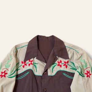 May include: A button-down shirt with a brown body and tan sleeves. The shirt features embroidered red flowers, green cacti, and teal accents. The collar and button placket are brown.