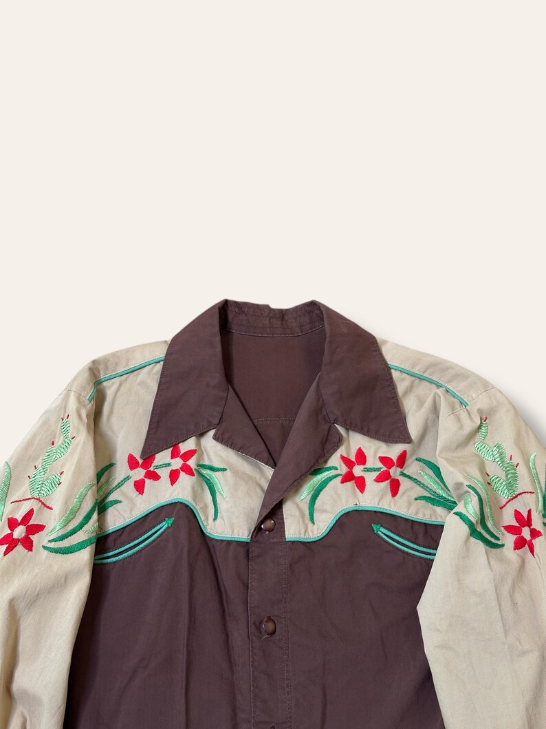 May include: A button-down shirt with a brown body and tan sleeves. The shirt features embroidered red flowers, green cacti, and teal accents. The collar and button placket are brown.