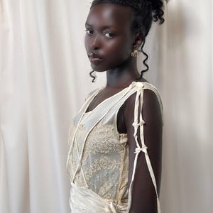 May include: A woman wearing a white, lace-trimmed dress with a tied sash and a V-neckline. The dress has thin straps that tie into bows on the arms.