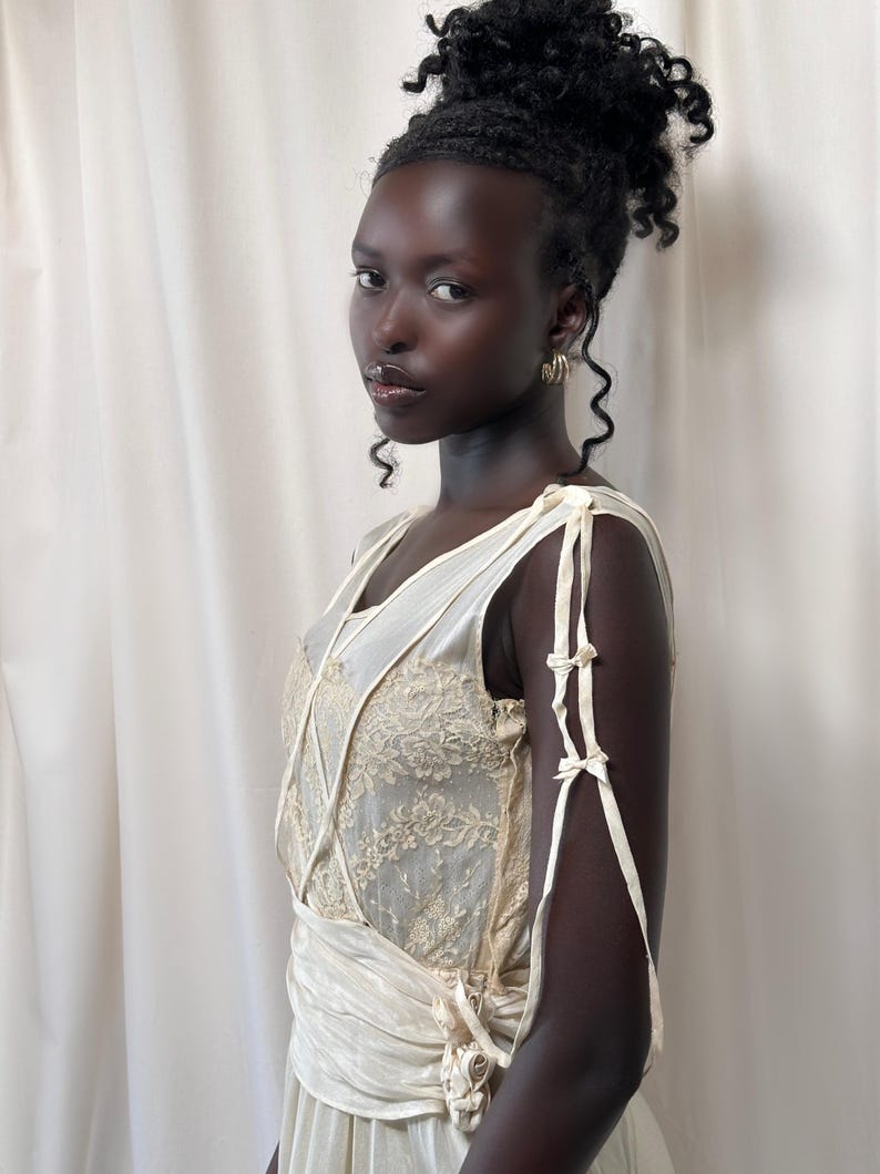 May include: A woman wearing a white, lace-trimmed dress with a tied sash and a V-neckline. The dress has thin straps that tie into bows on the arms.