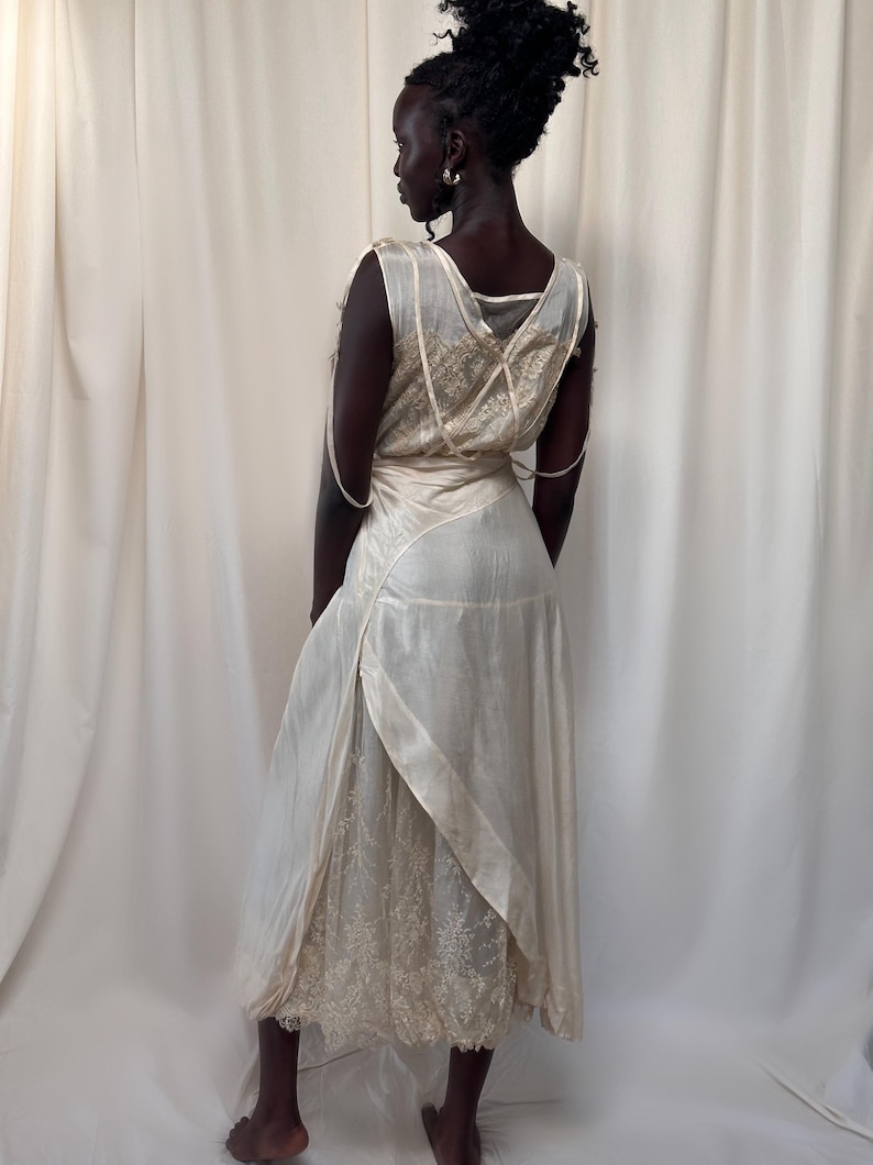 May include: A white vintage dress with a lace overlay and a satin sash. The dress has a sleeveless design and a flowing skirt.