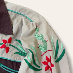 May include: Close-up of a tan shirt with brown collar and accents. The shirt features embroidered designs of red flowers, green cacti, and green leaves. The trim is a light teal color.
