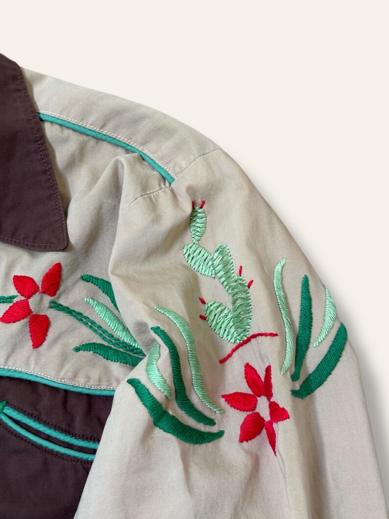 May include: Close-up of a tan shirt with brown collar and accents. The shirt features embroidered designs of red flowers, green cacti, and green leaves. The trim is a light teal color.