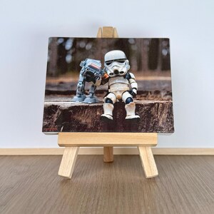 May include: A rectangular print on a small wooden easel featuring a white and black Stormtrooper figurine sitting next to a grey AT-ST walker toy. The figures are positioned on a textured brown log surface with a blurred forest background.