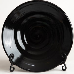 May include: A black ceramic plate with a glossy finish and a slightly raised center. The plate is on a black metal stand.