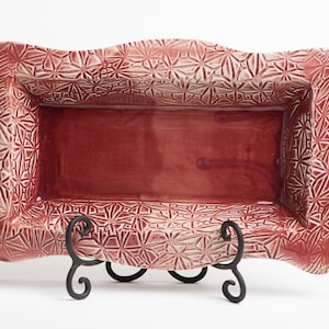 Pode incluir: Um prato de servir de cerâmica vermelha com um padrão decorativo em relevo. O prato está sobre um suporte de metal preto.