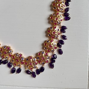 May include: Gold-toned necklace featuring floral pendants with red and purple accents. The necklace is adorned with dangling, teardrop-shaped, dark purple beads. The necklace is displayed on a white surface.