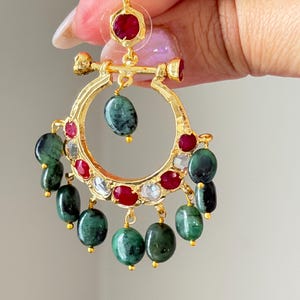 May include: Gold-toned hoop earrings with ruby and emerald accents. The earrings feature a circular design with dangling emerald beads and ruby and clear stone embellishments. The earrings are held up by a hand.