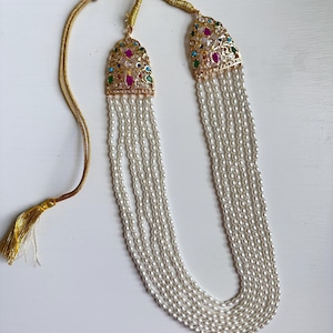 Op de afbeelding: Een goudkleurige ketting met meerdere strengen witte parels. De ketting heeft sierlijke gouden hangers versierd met kleurrijke edelstenen. De ketting heeft een kwastje aan het einde van het koord.