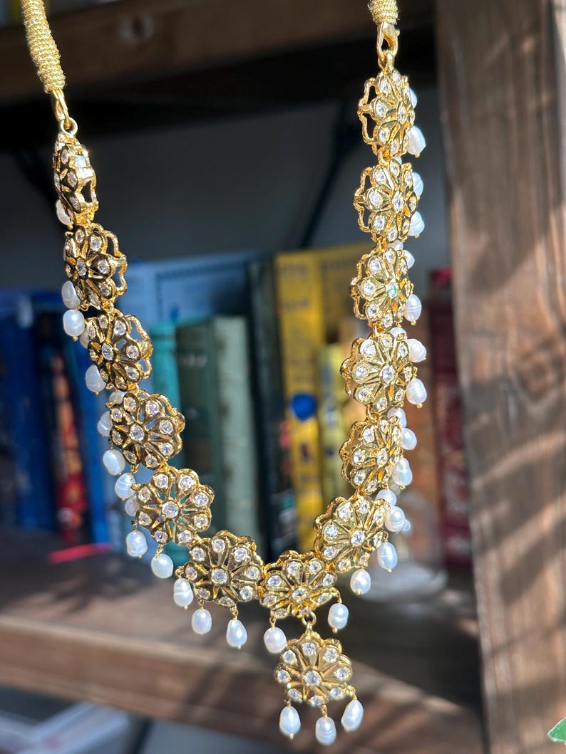 May include: A gold necklace with a floral design and white pearl accents. The necklace is made of metal and has a delicate, intricate design.