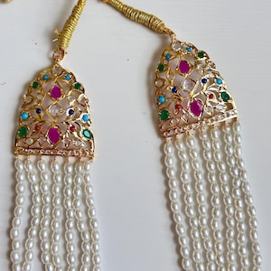 Op de afbeelding: Een goudkleurige ketting met een hanger met kleurrijke edelstenen en een parelstrengontwerp. De hanger heeft een filigraan patroon met rode, groene, blauwe en heldere stenen. De ketting heeft meerdere strengen kleine, witte parels.