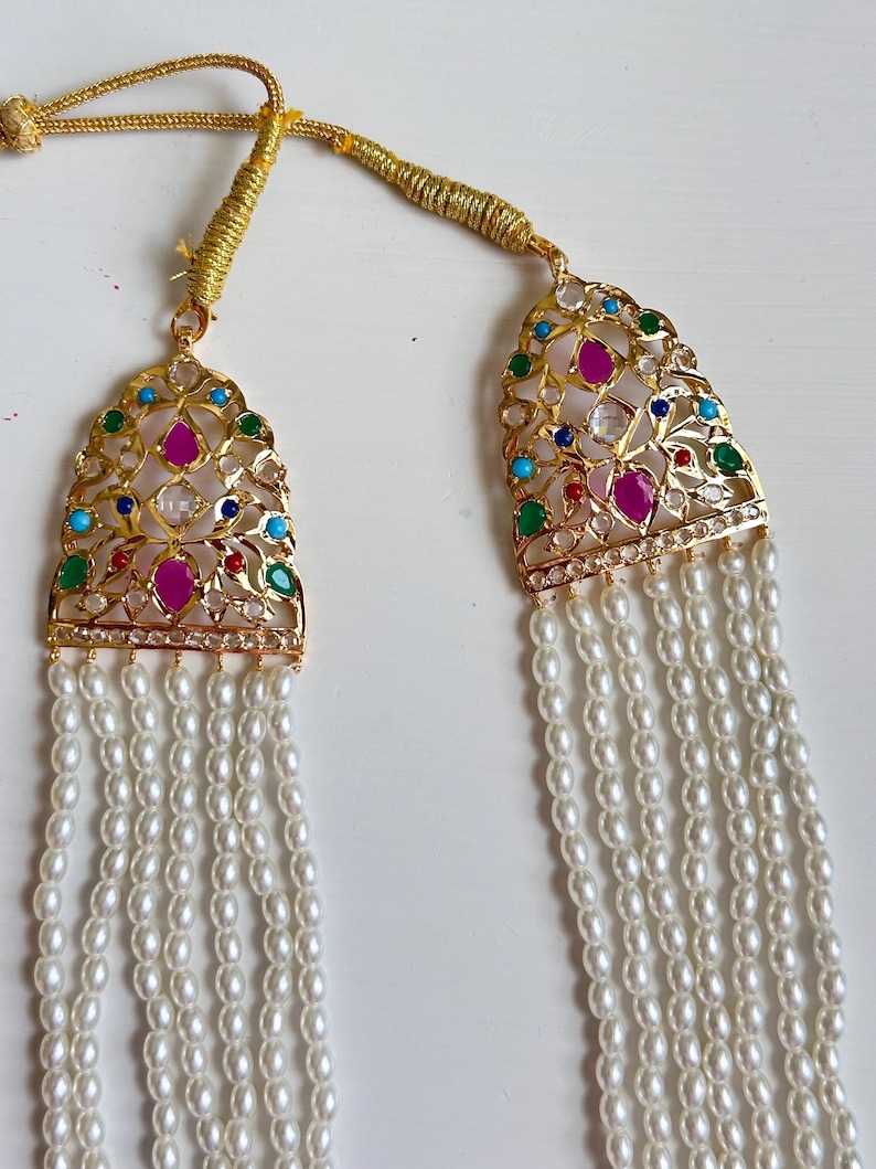 Op de afbeelding: Een goudkleurige ketting met een hanger met kleurrijke edelstenen en een parelstrengontwerp. De hanger heeft een filigraan patroon met rode, groene, blauwe en heldere stenen. De ketting heeft meerdere strengen kleine, witte parels.