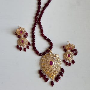 Pode incluir: Um conjunto de colar e brincos dourados com um pendente em forma de folha e brincos a condizer. O pendente e os brincos apresentam uma pedra central de cor rubi e pedras transparentes. O colar é feito de contas de cor grená.