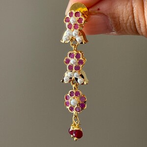 May include: A gold-colored earring with a floral design. The earring features red and white gemstones arranged in a flower shape, with a cluster of red gemstones at the bottom. The earring is held up by a person's fingers.