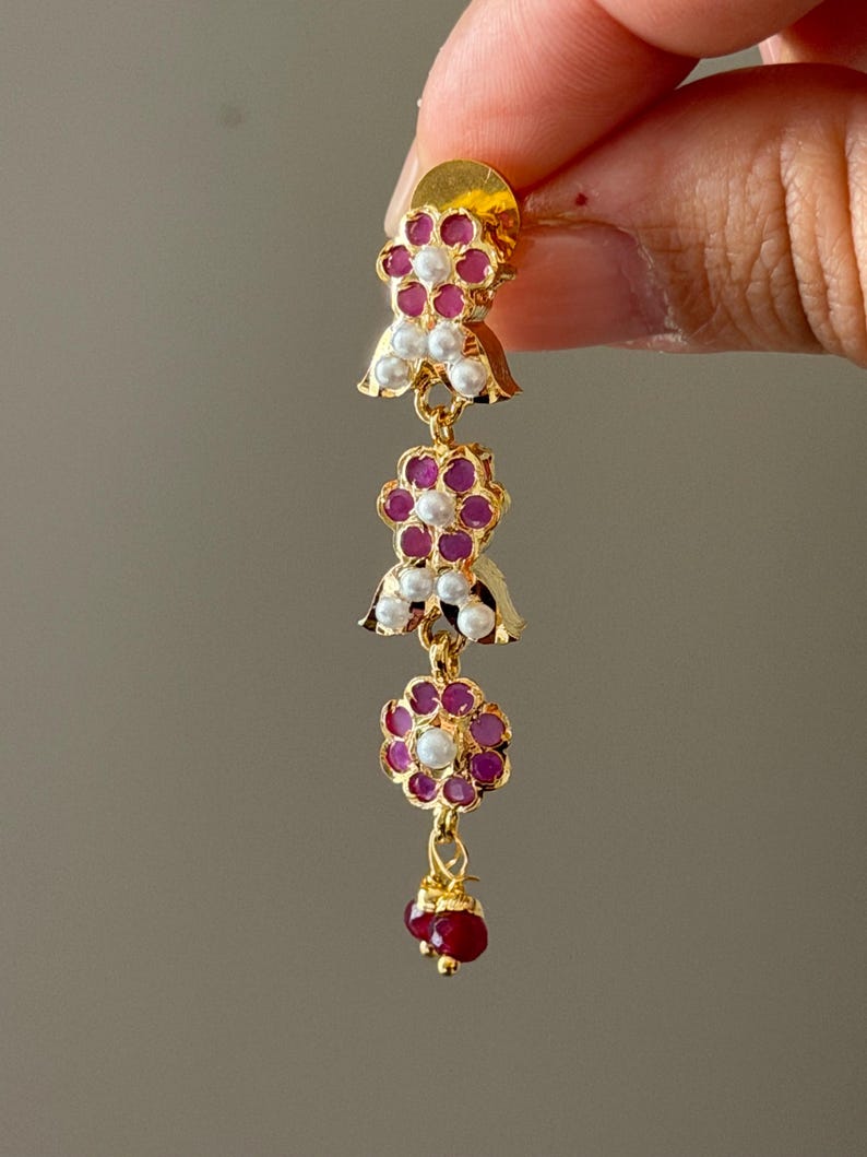 May include: A gold-colored earring with a floral design. The earring features red and white gemstones arranged in a flower shape, with a cluster of red gemstones at the bottom. The earring is held up by a person's fingers.