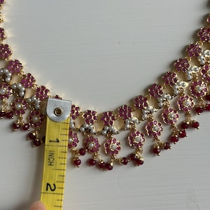 May include: Gold-toned necklace with ruby-colored floral accents and small white pearls. The necklace is approximately 2 inches in length, as measured by a yellow ruler.