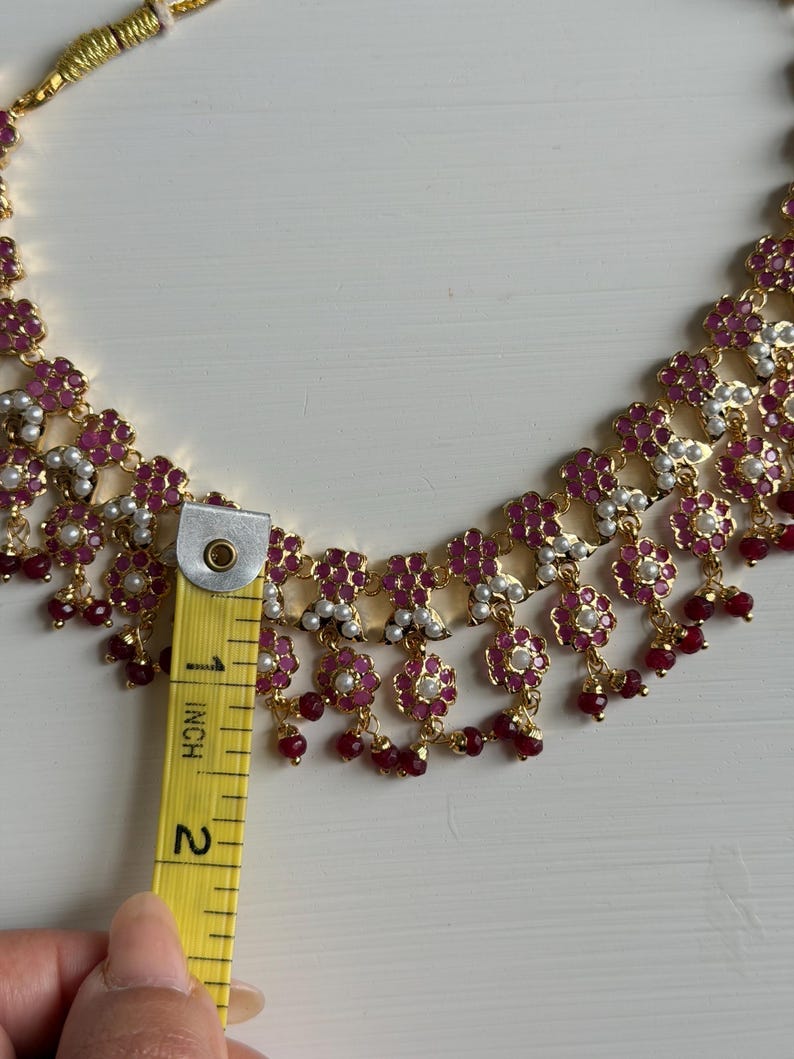 May include: Gold-toned necklace with ruby-colored floral accents and small white pearls. The necklace is approximately 2 inches in length, as measured by a yellow ruler.
