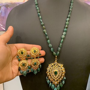 K&ouml;nnte beinhalten: Eine Halskette und Ohrringe aus gr&uuml;nem Smaragd mit einem goldenen Anh&auml;nger mit floralem Design und wei&szlig;en Edelsteinen.
