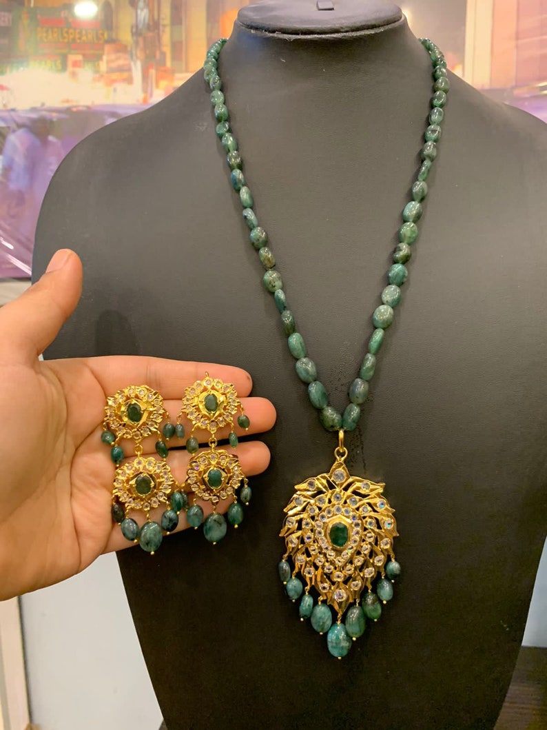 K&ouml;nnte beinhalten: Eine Halskette und Ohrringe aus gr&uuml;nem Smaragd mit einem goldenen Anh&auml;nger mit floralem Design und wei&szlig;en Edelsteinen.