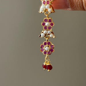 May include: Gold-toned earring with a cascading design. Features ruby-colored stones and white pearls set in floral and bell-shaped patterns. The earring ends with two ruby-colored beads. A delicate and ornate piece of jewelry.