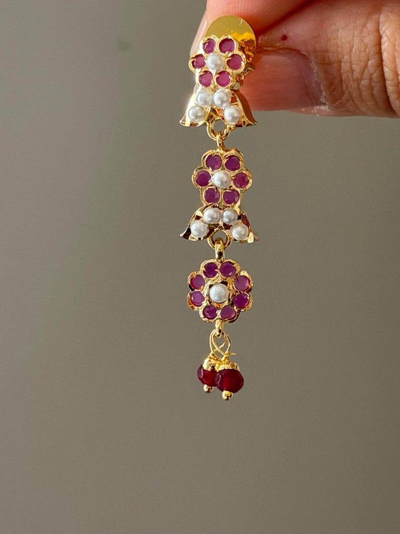 May include: Gold-toned earring with a cascading design. Features ruby-colored stones and white pearls set in floral and bell-shaped patterns. The earring ends with two ruby-colored beads. A delicate and ornate piece of jewelry.