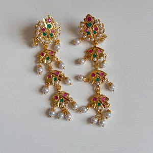 May include: A pair of gold-toned earrings with a floral design. Each earring features red, green, and white accents, with dangling faux pearls. The earrings are designed with multiple tiers and are displayed on a white surface.