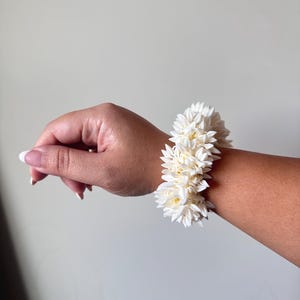 K&ouml;nnte beinhalten: Ein wei&szlig;es Blumenarmband aus kleinen, zarten Bl&uuml;ten. Das Armband wird am Handgelenk getragen.