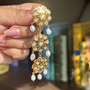May include: Gold dangle earrings with three flower-shaped charms and white pearl accents.