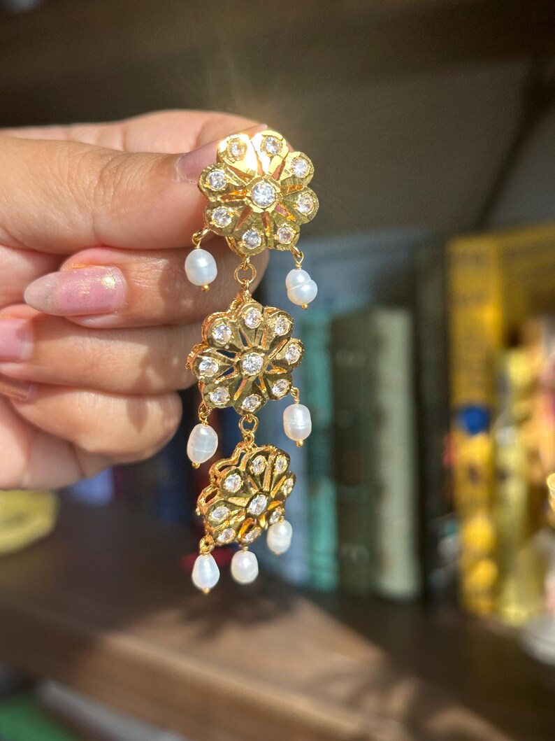 May include: Gold dangle earrings with three flower-shaped charms and white pearl accents.