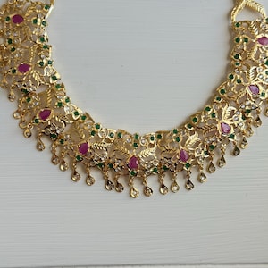 May include: A gold-toned necklace with intricate floral and leaf designs. The necklace is adorned with pink, green, and clear gemstones. Small teardrop-shaped pendants dangle from the bottom edge of the necklace.