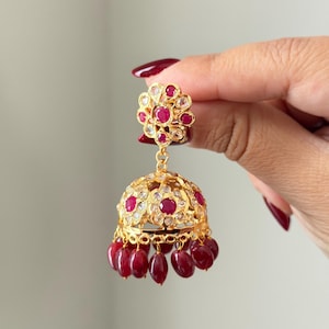 May include: An ornate gold-coloured earring with a floral top and a bell-shaped bottom. The earring is decorated with red and clear gemstones. Several teardrop-shaped red gems dangle from the bottom. The earring is held up by a hand.
