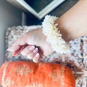 K&ouml;nnte beinhalten: Ein wei&szlig;es Blumenarmband aus kleinen wei&szlig;en Bl&uuml;ten. Das Armband wird am Handgelenk getragen.