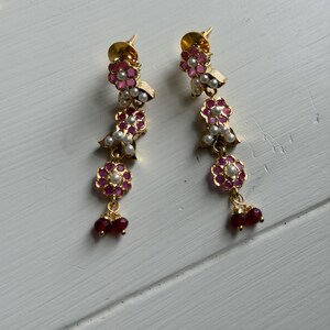 May include: A pair of gold-toned earrings with a floral design. Each earring features pink and white flower-shaped accents, small white pearls, and dangling red beads. The earrings are displayed on a white surface.